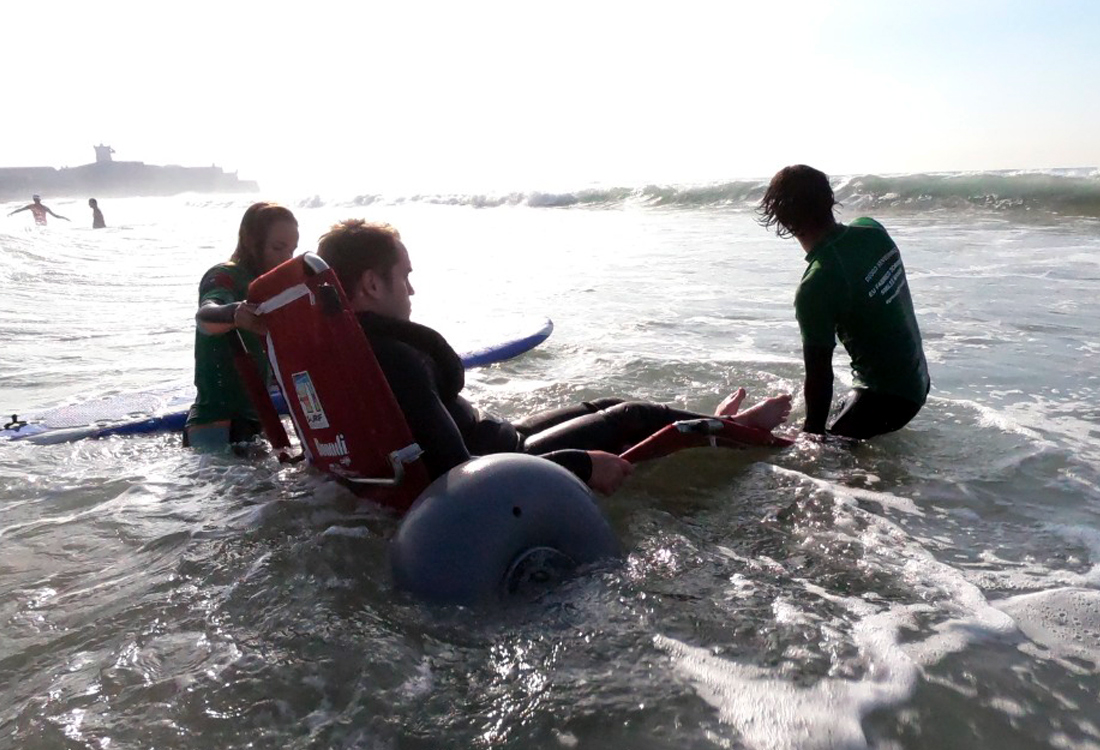 FFALP class of 2019 at surfing session with participant being transferred from a beach wheelchair to a surf board, with the support of two volunteers