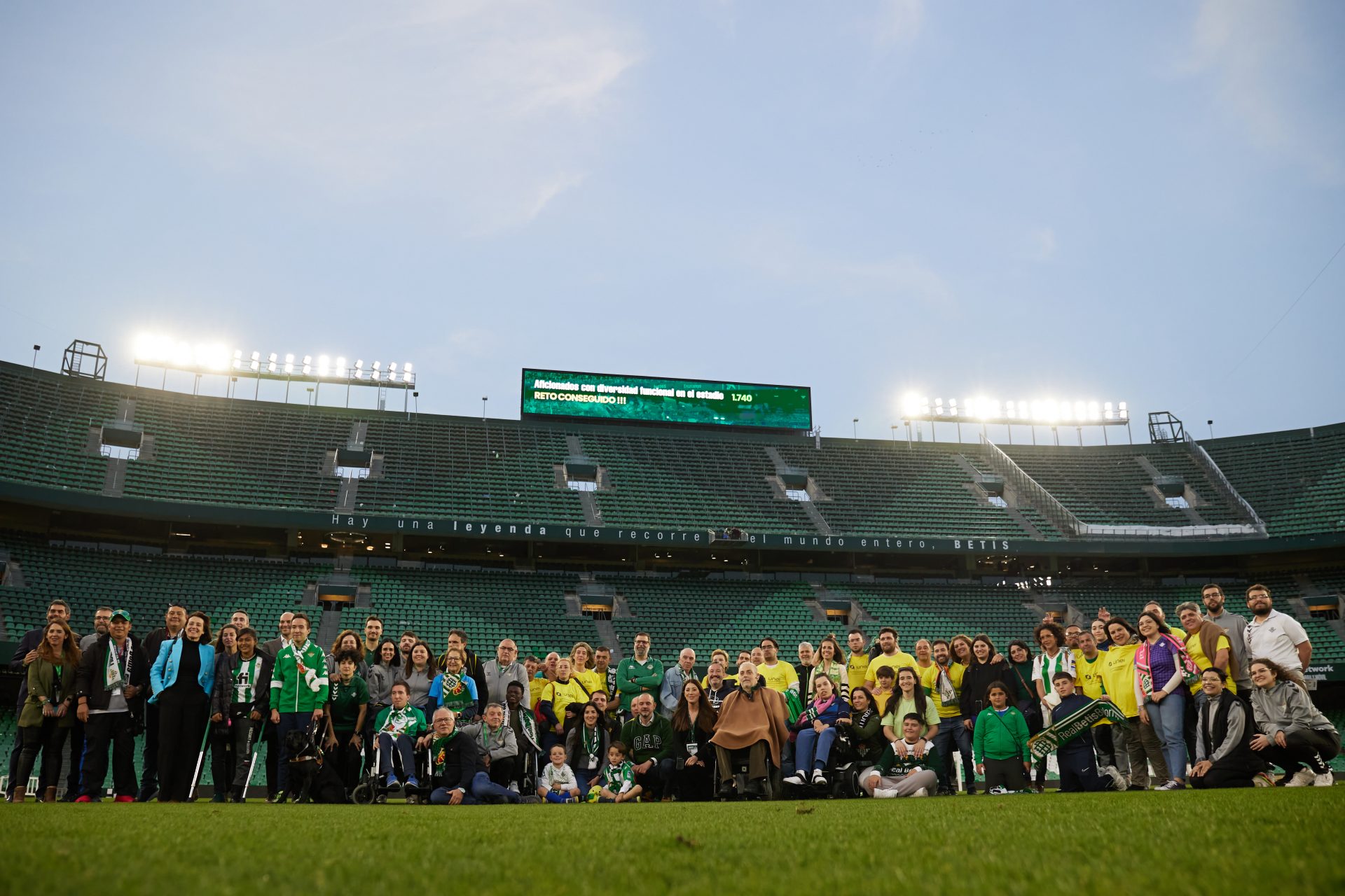 Group of disabled fans and organisers stands on the Real Betis Balompié pitch in the Benito Vilamarin Stadium after the match. In the backgroud empty stands and giant screen showing the record breaking number of disabled fans attending the match