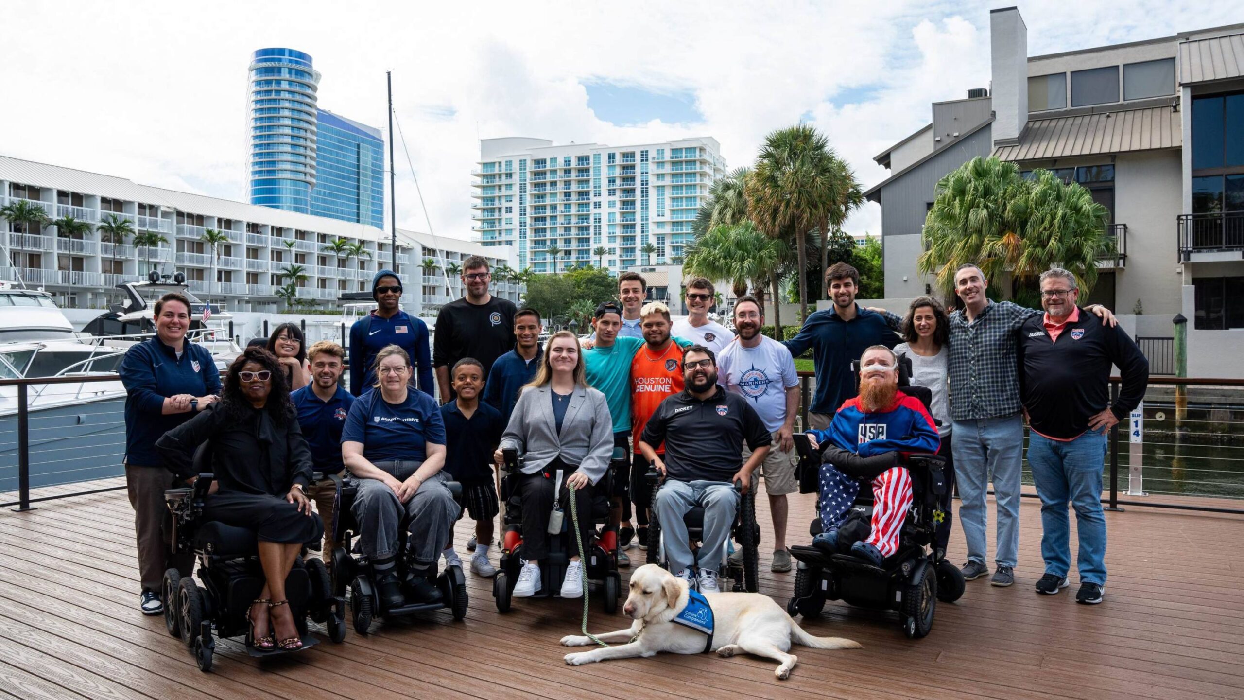 A group of approximately 20 people posing for a group photo on a wooden pier or boardwalk. The group is diverse in age and includes several individuals using power wheelchairs. In the center foreground, a cream-colored service dog lies on the deck. The people are dressed in a mix of casual and business-casual attire, including some wearing "USA" branded athletic gear. Behind the group is a marina with yachts, modern high-rise buildings, and palm trees under a bright, partly cloudy sky.