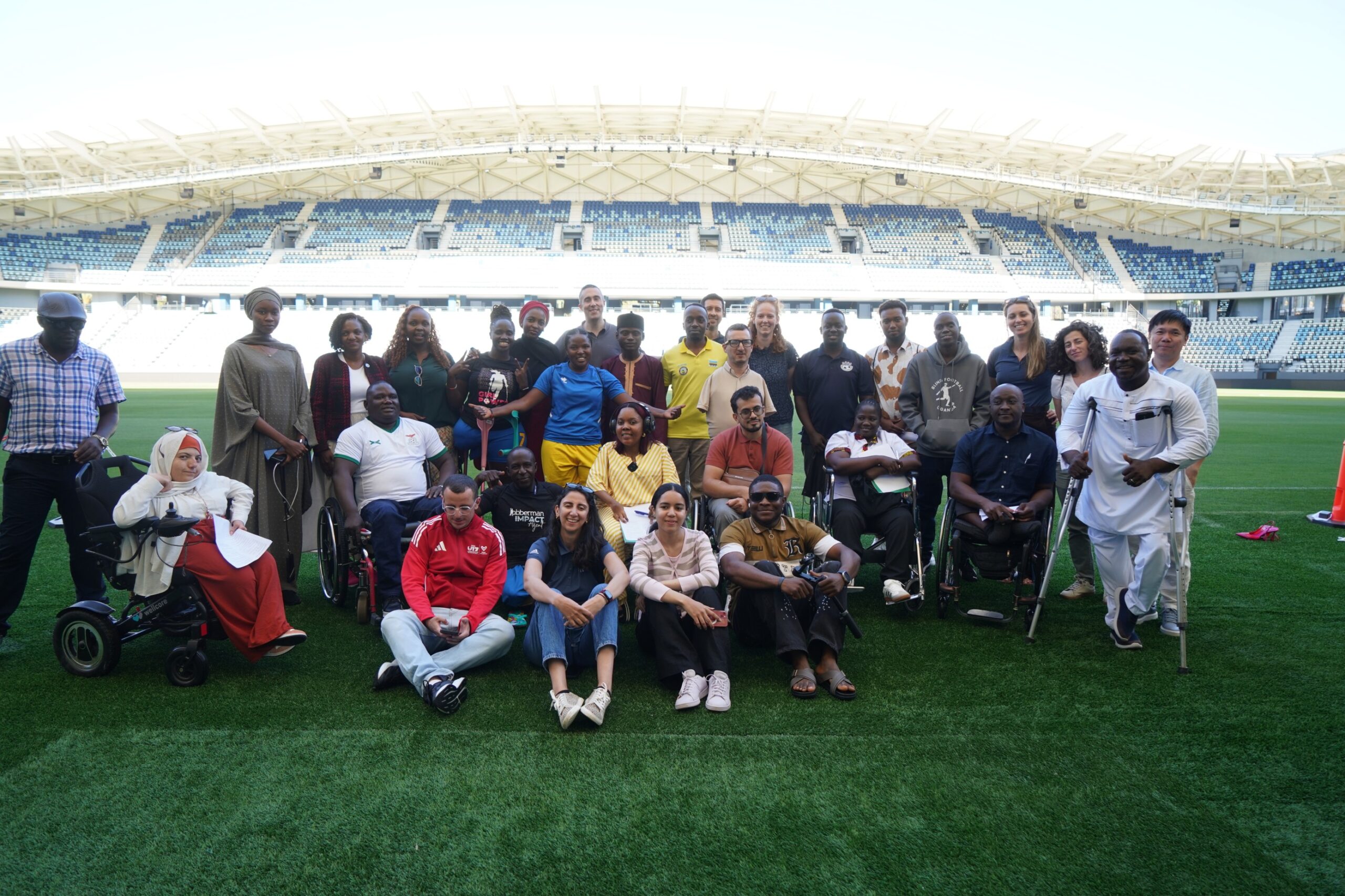 A large, diverse group of people poses for a commemorative photo on the bright green grass of a professional football stadium. The group includes individuals standing, sitting on the grass, and using various mobility aids such as wheelchairs and crutches. The empty blue and white stadium seats are visible in the background under a clear sky.