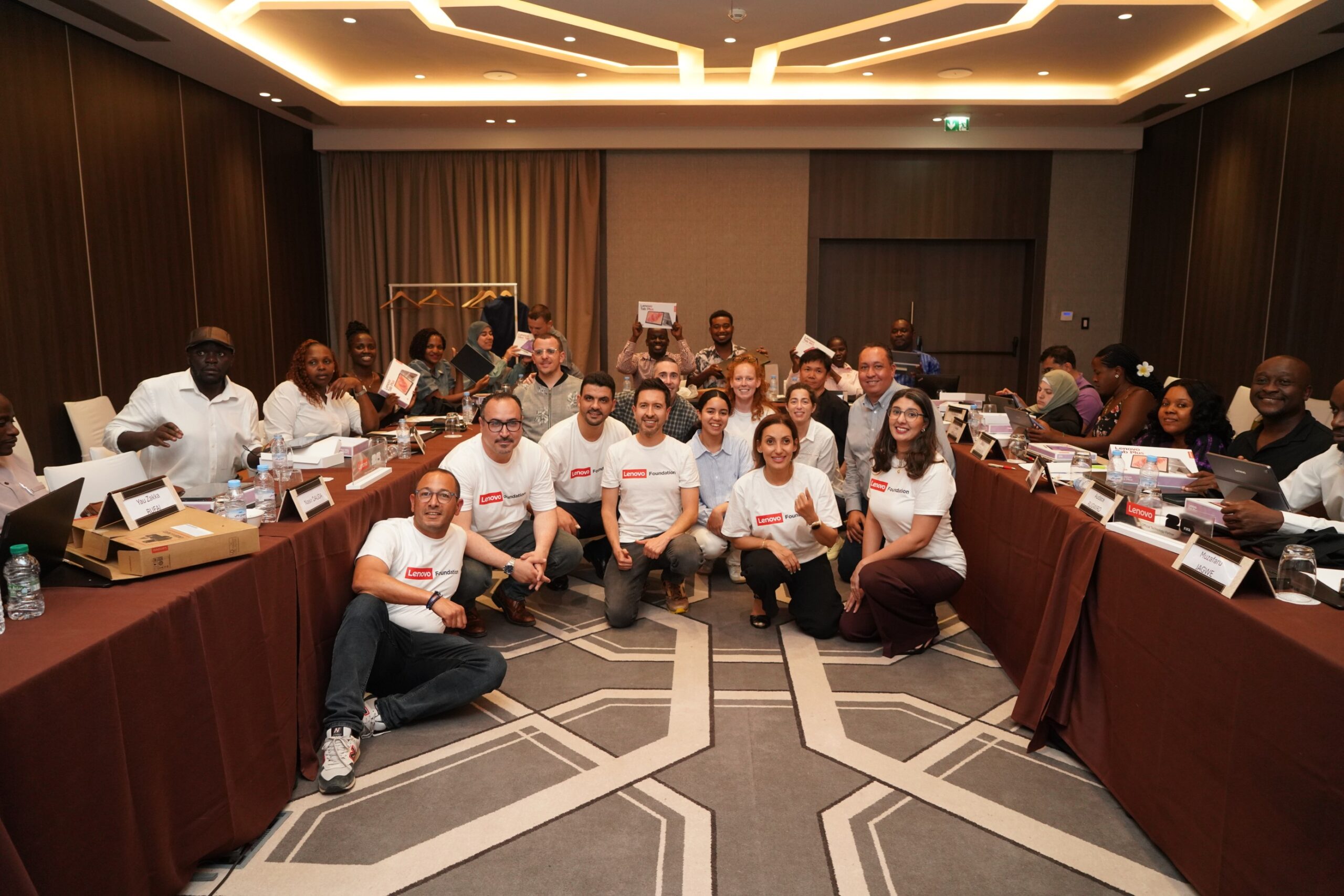 A large group of people poses in a U-shaped configuration within a modern conference room with geometric ceiling lights. In the front, five individuals are in a group, with four in white 'Lenovo Foundation' t-shirts and one person crouched; on the far left, a person winks, and a Lenovo box is on the table next to them. Behind the table, many attendees hold small white gift boxes, and place cards are visible.