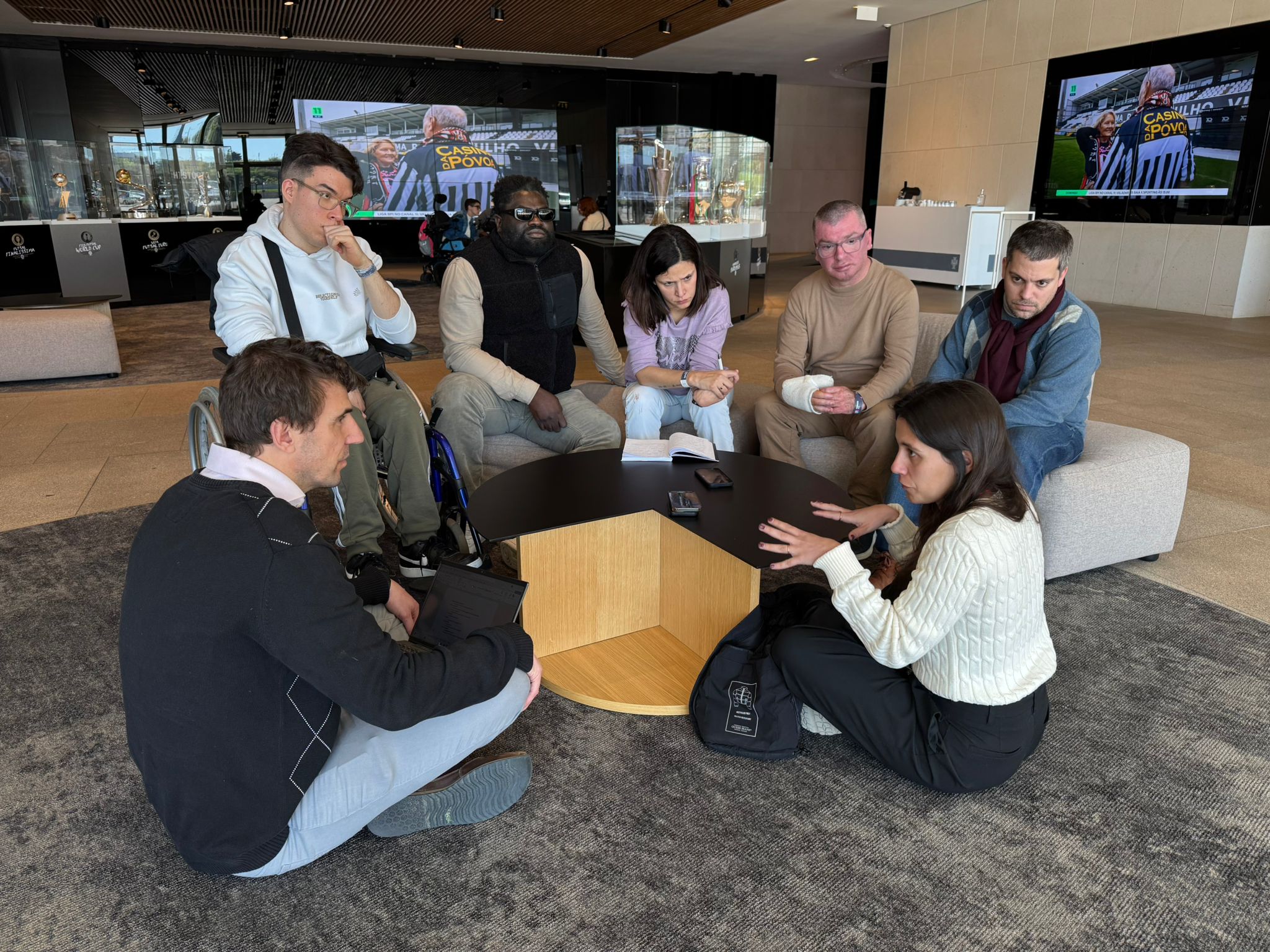 A high-angle, wide photograph shows seven diverse people gathered in an informal discussion circle on a patterned rug in a modern lounge. Two individuals are seated on a light grey ottoman sofa, and two others sit in low-slung, fabric-covered chairs with wooden frames and matching footrests. Three participants sit cross-legged on the floor, including a woman in a white knitted sweater who is speaking, gesturing with both hands towards the group. A man in a wheelchair is included in the circle, positioned between the ottoman and floor-sitters. In the center, there's a low-profile, multi-tiered black and wood coffee table with notebooks, smartphones, and papers on it. The background features floor-to-ceiling windows showing a blurred city street, and an exposed concrete wall with a few pieces of abstract art. The overall atmosphere is warm and collaborative.